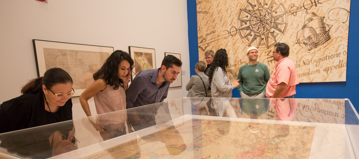 image of college students and members looking at cases with maps in them within museum gallery